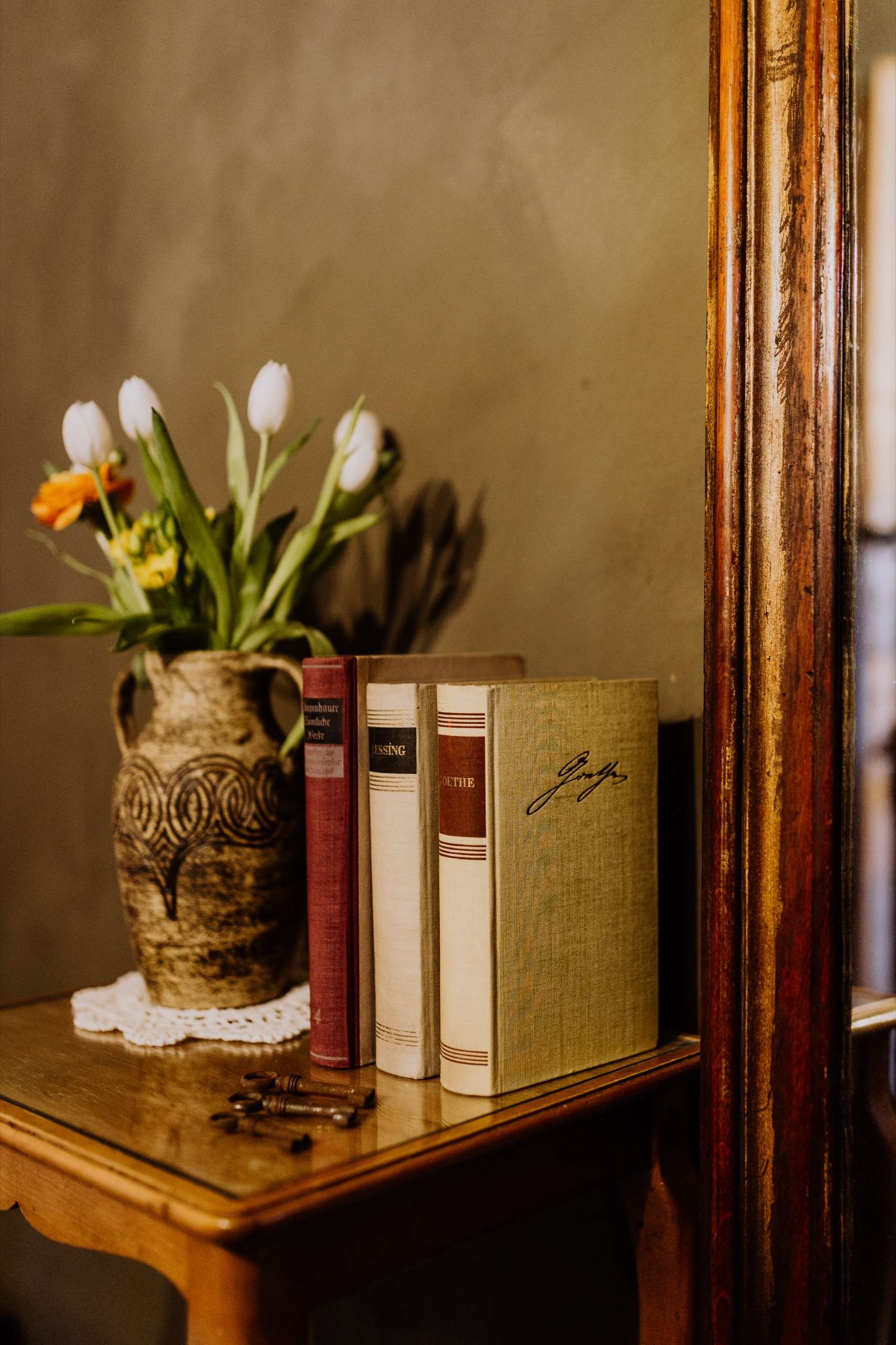 Frische Tulpen im Steingutkrug neben antiken Büchern und alten Schlüsseln vor dem Goldspiegel